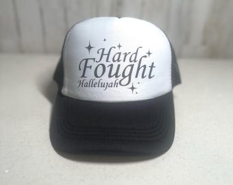 May include: A black and white trucker hat with the words "Hard Fought Hallelujah" in a script font. The hat has a black brim and a white front panel with the text and star accents. The mesh back is black.