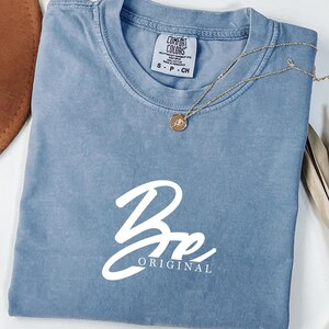 Może przedstawiać: Niebieska koszulka Comfort Colors z napisem "Be Original" w białym skrypcie. Złoty naszyjnik z wisiorkiem spoczywa na koszulce. Koszulka jest złożona, a brązowy kapelusz jest widoczny w lewym górnym rogu.