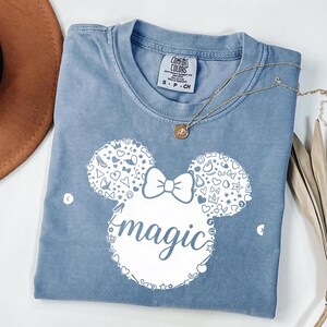 Puede incluir: Una camiseta azul Comfort Colors con un dise&ntilde;o blanco de Minnie Mouse y la palabra "magic". El dise&ntilde;o incluye un lazo y varias formas. Un collar dorado est&aacute; sobre la camiseta.