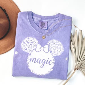 Puede incluir: Una camiseta Comfort Colors lavanda con un dise&ntilde;o blanco de cabeza de rat&oacute;n y la palabra "magic". La cabeza del rat&oacute;n est&aacute; adornada con corazones y estrellas. Tambi&eacute;n se ve un collar dorado y un sombrero marr&oacute;n.