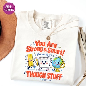 May include: A cream-coloured t-shirt featuring a graphic with the text "You Are Strong & Smart! You can do it! THOUGH STUFF" and cartoon illustrations of a yellow ruler, a white notepad, and a globe with faces, arms, and legs.