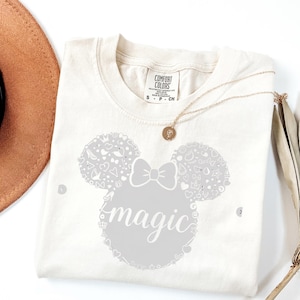 Puede incluir: Camiseta color crema con un gr&aacute;fico gris de una cabeza de rat&oacute;n, un lazo y la palabra "magic". La cabeza del rat&oacute;n est&aacute; llena de peque&ntilde;as formas decorativas. Un collar dorado con un colgante descansa sobre la camisa.