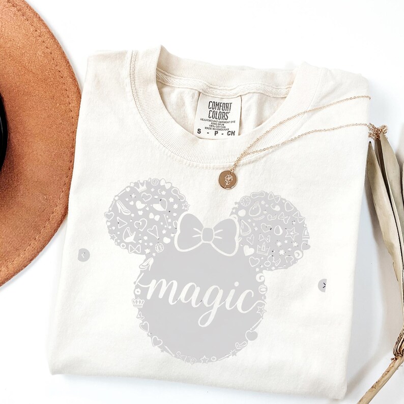 Puede incluir: Camiseta color crema con un gr&aacute;fico gris de una cabeza de rat&oacute;n, un lazo y la palabra "magic". La cabeza del rat&oacute;n est&aacute; llena de peque&ntilde;as formas decorativas. Un collar dorado con un colgante descansa sobre la camisa.