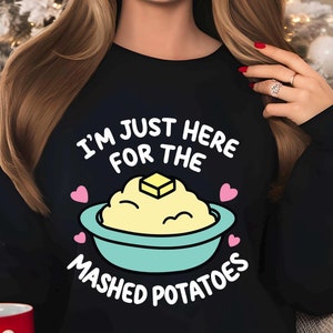 May include: Black sweatshirt with the text "I'M JUST HERE FOR THE MASHED POTATOES" in white. The graphic features a bowl of mashed potatoes with a pat of butter and pink hearts. A festive and humorous design.