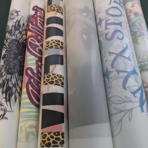 Puede incluir: Una colecci&oacute;n de hojas de papel blancas enrolladas con varios dise&ntilde;os. Los dise&ntilde;os incluyen un girasol en blanco y negro, texto en morado y rojo, un estampado de leopardo, una imagen en escala de grises de un ojo y texto floral azul.