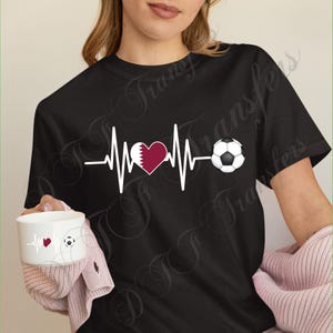 Puede incluir: Camiseta negra con un dise&ntilde;o de latido de coraz&oacute;n blanco, un coraz&oacute;n granate y un bal&oacute;n de f&uacute;tbol. El dise&ntilde;o tambi&eacute;n incluye la bandera de Qatar. Tambi&eacute;n se ve una taza blanca con un dise&ntilde;o similar.