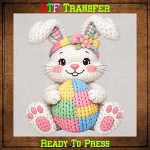 May include: A white knitted bunny holding a colorful patchwork egg. The bunny has pink cheeks, a flower on its head, and pink paw pads. The image includes the text "DTF TRANSFER" and "READY TO PRESS".