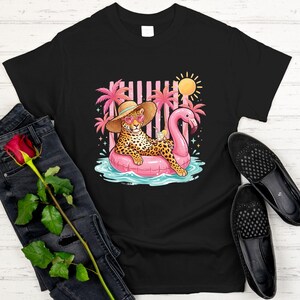 May include: Black t-shirt featuring a leopard wearing sunglasses and a straw hat, lounging on a pink flamingo float in a pool. The graphic includes a sun, palm trees, and a cocktail. A pair of black loafers and jeans are next to the shirt.