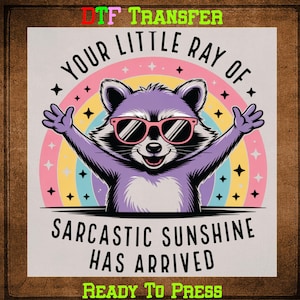 May include: A DTF transfer featuring a purple raccoon wearing pink sunglasses with arms outstretched in front of a rainbow. Text reads "YOUR LITTLE RAY OF SARCASTIC SUNSHINE HAS ARRIVED" and "READY TO PRESS".