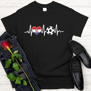 May include: Black t-shirt featuring a white heartbeat design with a Croatian flag heart and a soccer ball. The shirt is paired with dark denim jeans, a red rose, and black loafers. A casual and sporty theme.