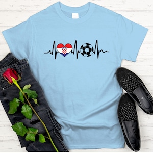 May include: Light blue t-shirt featuring a heartbeat design with a Croatian flag heart and a soccer ball. The shirt is displayed with black jeans, a red rose, and black shoes.