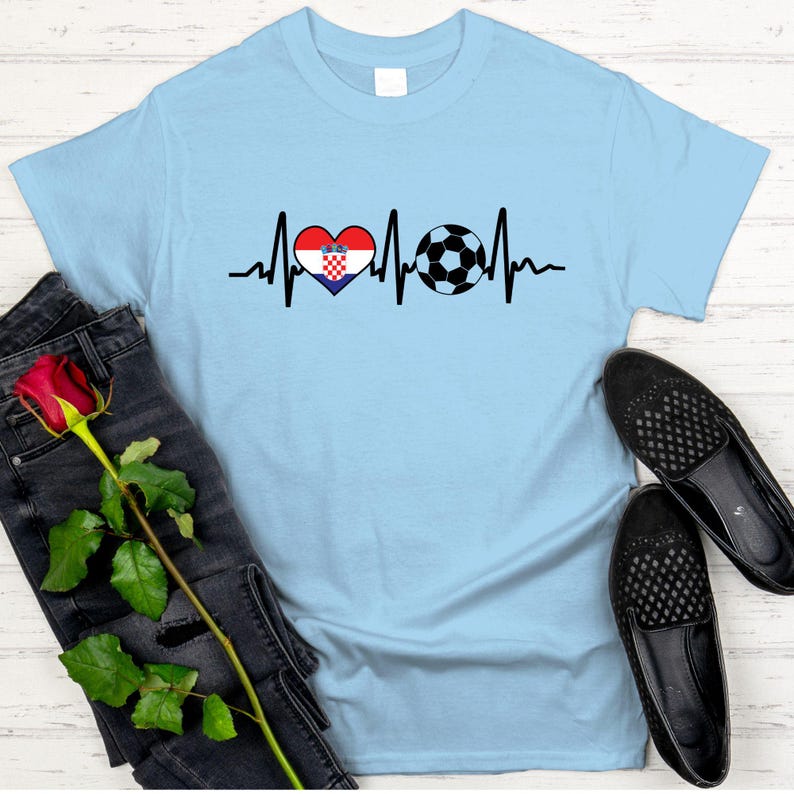 May include: Light blue t-shirt featuring a heartbeat design with a Croatian flag heart and a soccer ball. The shirt is displayed with black jeans, a red rose, and black shoes.