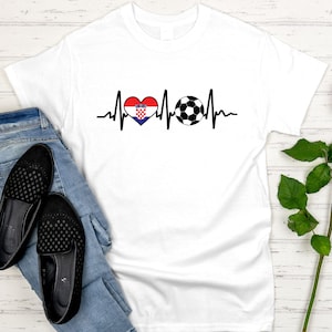 May include: White t-shirt featuring a black heartbeat line design with a heart in the colors of the Croatian flag and a soccer ball. The shirt is paired with blue jeans and black flats.