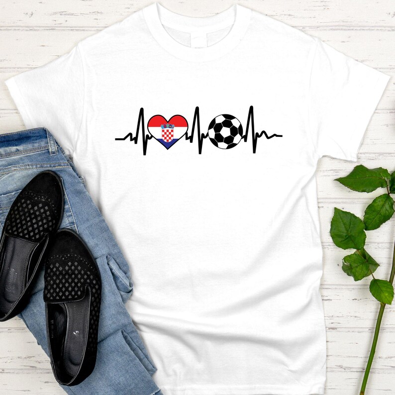 May include: White t-shirt featuring a black heartbeat line design with a heart in the colors of the Croatian flag and a soccer ball. The shirt is paired with blue jeans and black flats.
