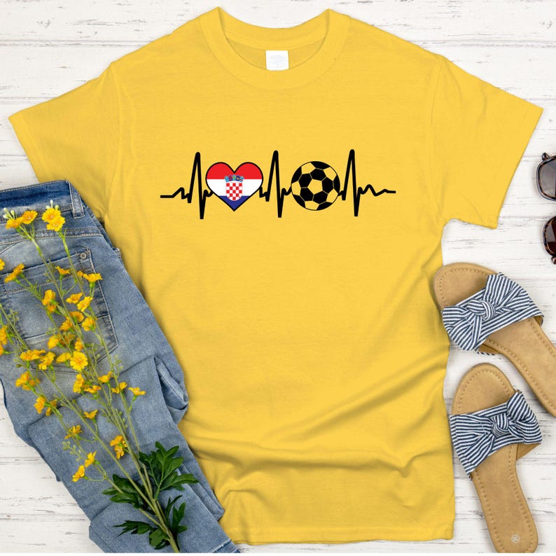 May include: A yellow t-shirt featuring a heartbeat design with a Croatian flag heart and a soccer ball. The shirt is displayed with blue jeans and sandals.