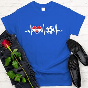 May include: A royal blue t-shirt featuring a white graphic of a heartbeat line, a heart with the Croatian flag design, and a soccer ball. The shirt is paired with black jeans and black shoes.
