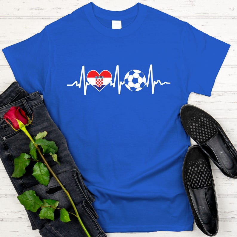 May include: A royal blue t-shirt featuring a white graphic of a heartbeat line, a heart with the Croatian flag design, and a soccer ball. The shirt is paired with black jeans and black shoes.