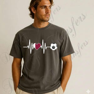 Puede incluir: Camiseta gris oscuro con un dise&ntilde;o de latido de coraz&oacute;n blanco, un coraz&oacute;n granate con la bandera de Qatar y un bal&oacute;n de f&uacute;tbol. La camiseta de manga corta est&aacute; hecha de un material suave.