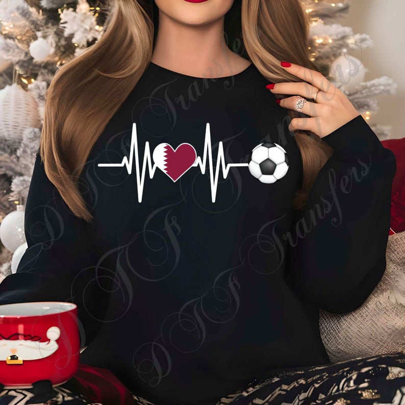 Puede incluir: Sudadera negra con un dise&ntilde;o de latido de coraz&oacute;n blanco. El dise&ntilde;o incluye un coraz&oacute;n granate con la bandera de Qatar y un bal&oacute;n de f&uacute;tbol. La sudadera es de color negro liso. El dise&ntilde;o est&aacute; centrado en el pecho.