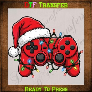 May include: A red video game controller wearing a red and white Santa hat, wrapped in a string of colorful Christmas lights. The text DTF TRANSFER READY TO PRESS is visible at the top and bottom of the graphic design on a light gray background.