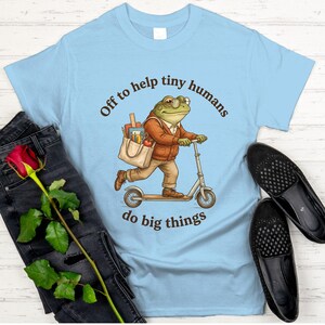 May include: Light blue t-shirt with a whimsical illustration of a frog riding a scooter, wearing glasses and carrying a tote bag. The text on the shirt reads "Off to help tiny humans do big things."