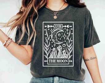 May include: Dark gray t-shirt featuring a white graphic of a crescent moon, stars, and crystals, with the text "THE MOON" and the Roman numeral "XVIII". The design is framed within a rectangular border.