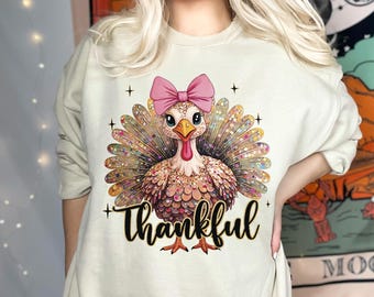 May include: Beige sweatshirt featuring a colorful turkey illustration with a pink bow and the word "Thankful" in black script. The turkey has a detailed feather design and is surrounded by small black crosses.