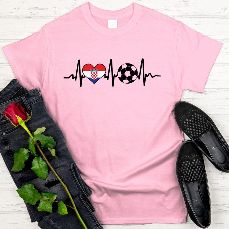 May include: A light pink t-shirt featuring a black heartbeat line design. The design includes a heart with the Croatian flag and a soccer ball. The shirt is paired with black jeans and black shoes.