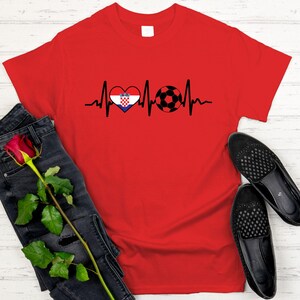 May include: A red t-shirt featuring a black heartbeat line design with a heart containing the Croatian flag and a soccer ball. The shirt is paired with black jeans, a red rose, and black loafers.