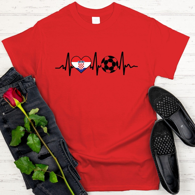May include: A red t-shirt featuring a black heartbeat line design with a heart containing the Croatian flag and a soccer ball. The shirt is paired with black jeans, a red rose, and black loafers.