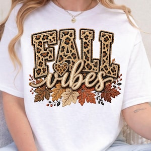 May include: A white t-shirt featuring the text FALL in large block letters with a brown and black leopard print pattern. Below, the word vibes is written in a cursive script, accompanied by a small leopard print heart and various autumn leaves.
