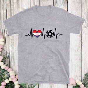 May include: A heather gray t-shirt featuring a graphic design. The design includes a heart with the Croatian flag, a soccer ball, and a heartbeat line. The shirt is a short-sleeved crew neck.