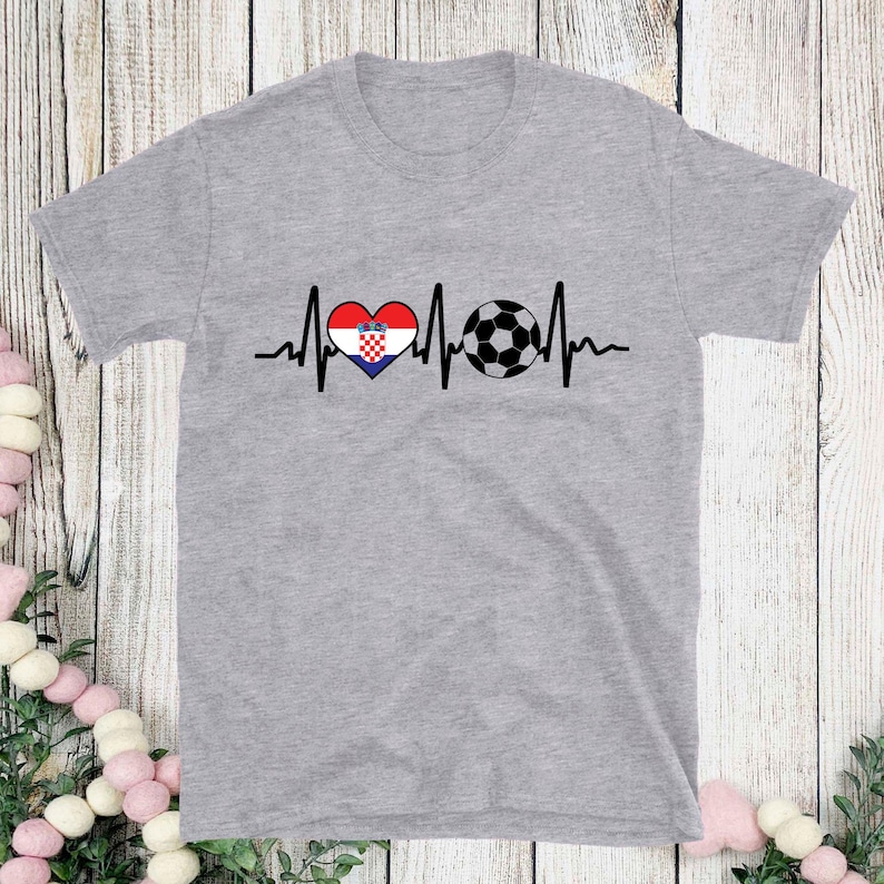 May include: A heather gray t-shirt featuring a graphic design. The design includes a heart with the Croatian flag, a soccer ball, and a heartbeat line. The shirt is a short-sleeved crew neck.