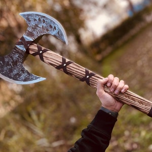 Könnte beinhalten: Eine Doppelkopf-Axt mit einem verwitterten, strukturierten Stahlkopf und einem Holzgriff. Der Axtkopf hat einen blau-grauen Farbton, und der Griff ist mit braunen Lederriemen umwickelt. Der Holzgriff hat ein geschnitztes Design.