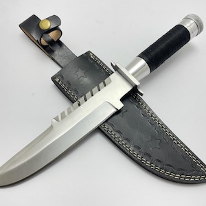May include: A stainless steel survival knife with a serrated blade and black wrapped handle rests in a black leather sheath. The sheath features star-shaped cutouts and white stitching, secured by a leather strap.