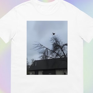 Könnte beinhalten: Weißes T-Shirt mit einem Foto eines Baumes und eines Vogels vor einem bewölkten Himmel. Ein Gebäude mit dunklem Dach ist unter dem Baum zu sehen. Das T-Shirt besteht aus einem weichen Material.