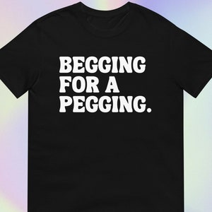 Könnte beinhalten: Schwarzes T-Shirt mit dem weißen Text "BEGGING FOR A PEGGING." Der Text ist fett und ohne Serifen. Das T-Shirt wird auf einem Farbverlauf-Hintergrund präsentiert.