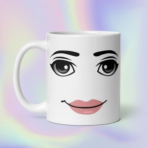 Può includere: Una tazza in ceramica bianca con una stampa grafica di un volto stilizzato con grandi occhi neri, sopracciglia scure arcuate e labbra rosa. Il design è centrato sulla parte anteriore della tazza, dotata di un manico curvo sul lato.