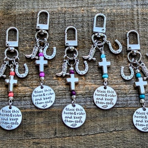 May include: Keychains with silver-tone charms, including crosses, horseshoes, and cowboy boots. Each keychain has a round charm with the text "Bless this horse & rider and keep them safe". The keychains are on a wooden surface.