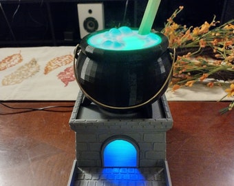 May include: A black cauldron with a green liquid and a straw sits on a gray stone base with a blue light. The base has a small archway with a blue light shining through.