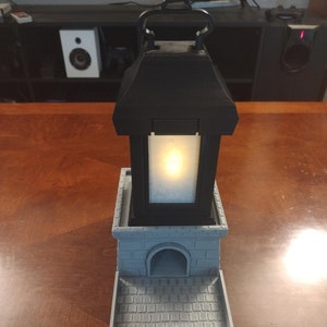 May include: A gray 3D printed dice tower with a black lantern on top. The lantern has a white light inside. The tower has a small archway on the front and a square platform on top.