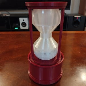 Könnte beinhalten: Eine rot-weiße Sanduhr mit zylindrischem Sockel und Oberteil, verbunden durch vertikale Stäbe. Die Sanduhrform ist mit einer weißen Substanz gefüllt. Der Artikel besteht aus Kunststoff und steht auf einer Holzoberfläche.