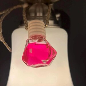 May include: A small, clear, faceted glass bottle filled with bright pink liquid, suspended from a light fixture. The bottle has a wooden cap and is hanging by a thin, woven cord. The background is a soft, white light.