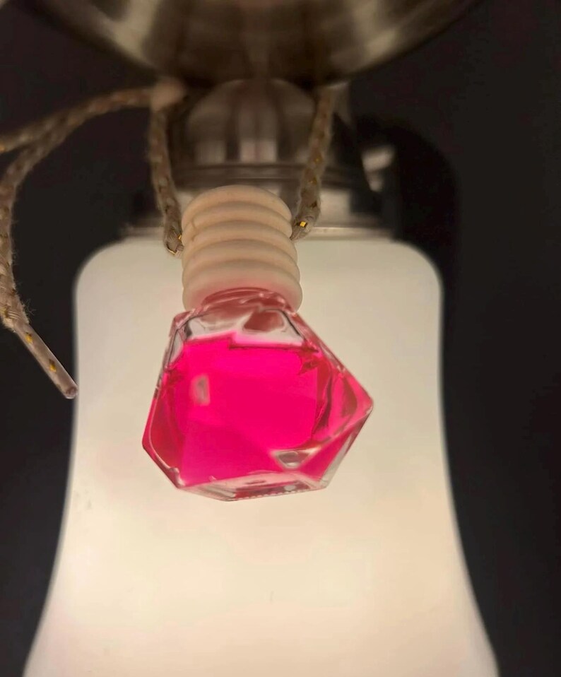 May include: A small, clear, faceted glass bottle filled with bright pink liquid, suspended from a light fixture. The bottle has a wooden cap and is hanging by a thin, woven cord. The background is a soft, white light.