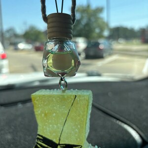 May include: A car air freshener, comprising a clear, geometric bottle with yellow liquid and a wooden cap, suspended by a string. Below, a yellow boot-shaped charm with black and red details hangs from a silver loop.