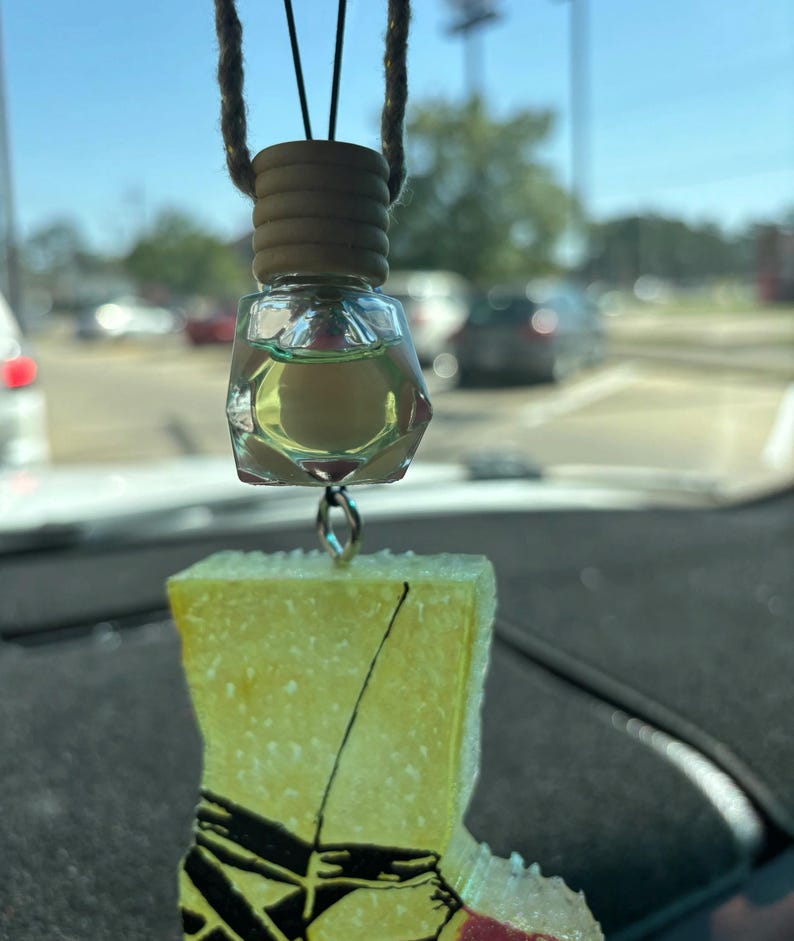 May include: A car air freshener, comprising a clear, geometric bottle with yellow liquid and a wooden cap, suspended by a string. Below, a yellow boot-shaped charm with black and red details hangs from a silver loop.