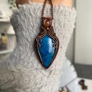 Pode incluir: Um pingente de pedra azul em forma de gota, envolto em um intrincado fio de cobre. O pingente apresenta uma pequena pedra redonda marrom-alaranjada acima da gema azul principal, suspenso por uma corrente de cobre correspondente.