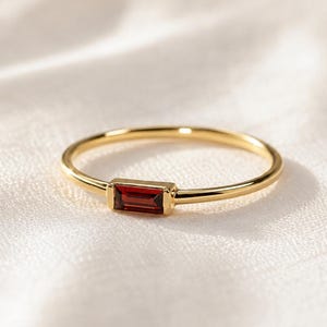 Könnte beinhalten: Ein dünner Goldring mit einem einzelnen, rechteckigen, im Baguetteschliff geschliffenen, tiefroten Granat-Edelstein, der horizontal in einer glatten Metallfassung sitzt. Der Ring hat eine polierte Oberfläche und ein schlichtes, schmales Design.
