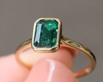Op de afbeelding: Een gouden ring met een rechthoekige, smaragdgeslepen groene edelsteen. De ring heeft een dunne, gouden band en een bezel setting die de smaragd omlijst. De ring wordt tegen een neutrale achtergrond gehouden.