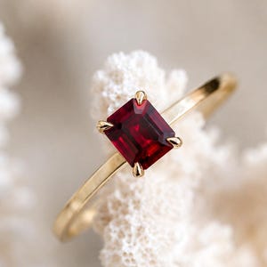 May include: A gold ring featuring a single rectangular emerald-cut deep red gemstone set in a four-prong setting. The band is thin and polished, showcasing a minimalist design that highlights the vibrant color and clarity of the central stone.